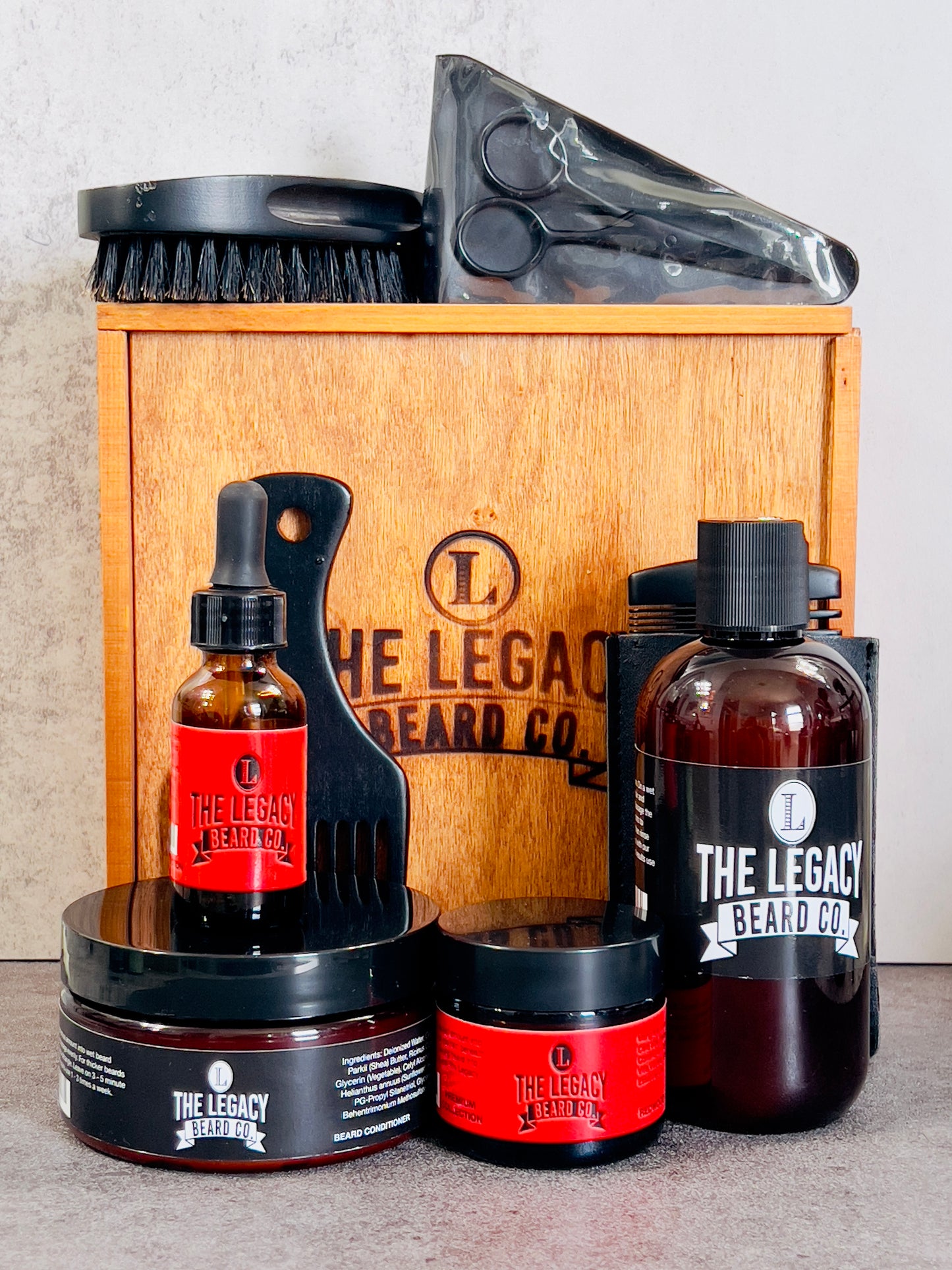 A grooming kit for beard care containing bottles of beard wash, conditioner, and oil, along with a beard butter container, wooden comb, boar bristle brush, beard pick, and stainless steel scissors, all displayed in front of a wooden box with the brand logo.