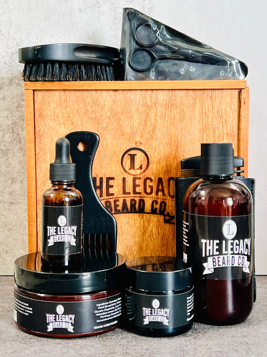 Beard kit with a white background on a concrete surface. This Kit includes:8 oz. Beard Wash, 8 oz., Beard Conditioner, 3 oz. Beard Enhancer, 1 oz. Beard Oil, scissors, beard brush, wooden comb, Wood keepsake box
