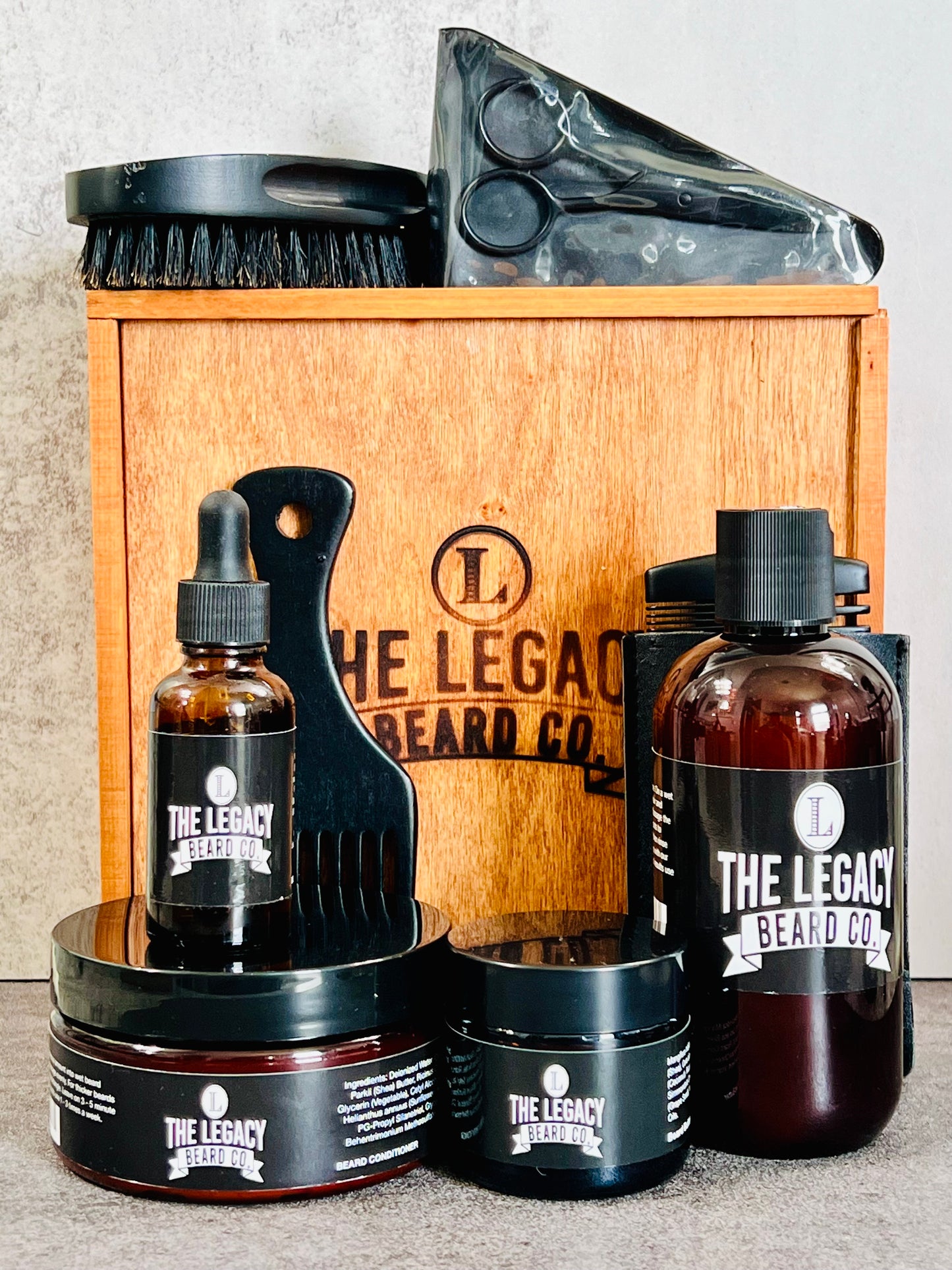 Beard kit with a white background on a concrete surface. This Kit includes:8 oz. Beard Wash, 8 oz., Beard Conditioner, 3 oz. Beard Enhancer, 1 oz. Beard Oil, scissors, beard brush, wooden comb, Wood keepsake box