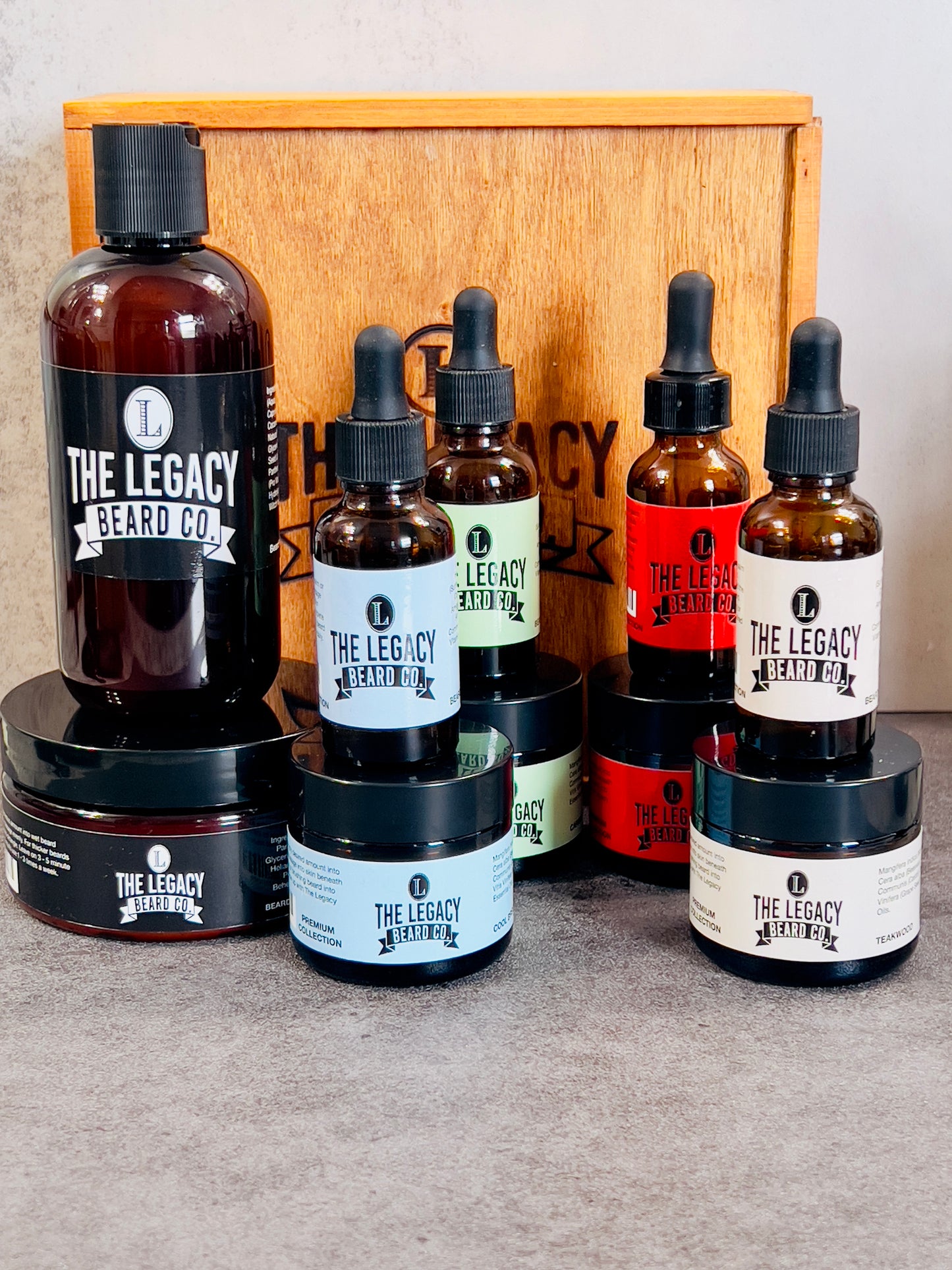 A collection of beard grooming products from The Legacy Beard Co., including beard wash, conditioner, butter, oil, all displayed in front of a wooden box.