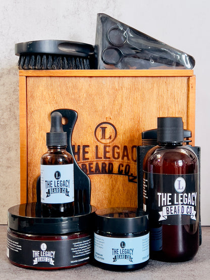 A grooming kit for beard care containing bottles of beard wash, conditioner, and oil, along with a beard butter container, wooden comb, boar bristle brush, beard pick, and stainless steel scissors, all displayed in front of a wooden box with the brand logo.