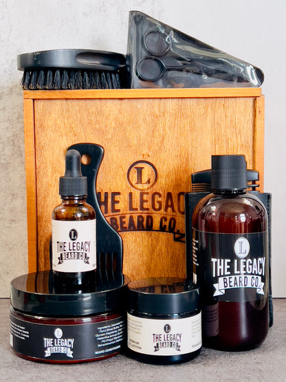 A grooming kit for beard care containing bottles of beard wash, conditioner, and oil, along with a beard butter container, wooden comb, boar bristle brush, beard pick, and stainless steel scissors, all displayed in front of a wooden box with the brand logo.