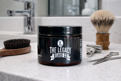 Black jar with 'The Legacy Beard Co.' logo on a bathroom counter with grooming tools.